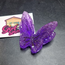 Load image into Gallery viewer, A purple glitter butterfly ornament or figurine displayed on a dark textured surface next to a small white card with a pink and yellow logo.
